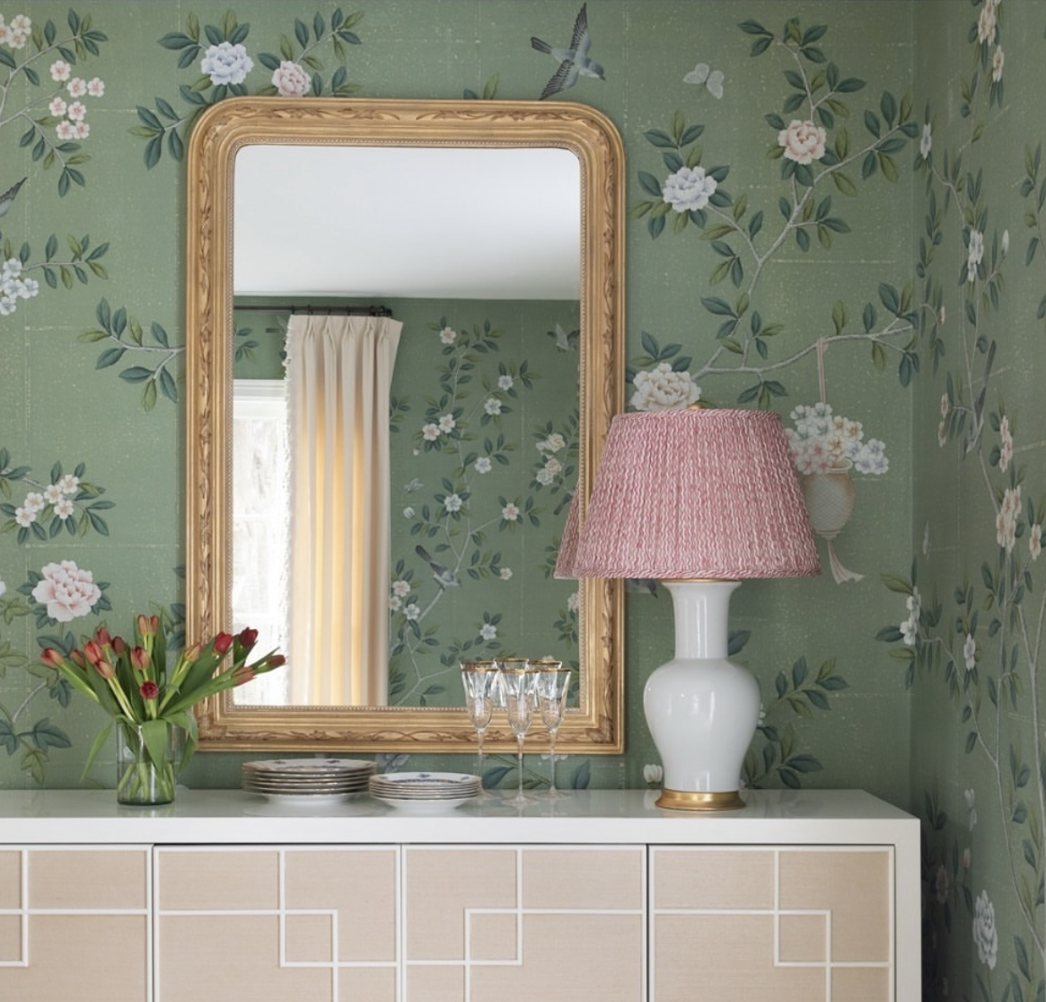 Chinoiserie hand-painted wallpaper 'Jade Garden' on antique green tea paper featuring white and pink blossoms with birds, styled with a white lacquer sideboard, a gold-framed mirror, red tulips in a glass vase, and a ceramic lamp with a red pleated shade.