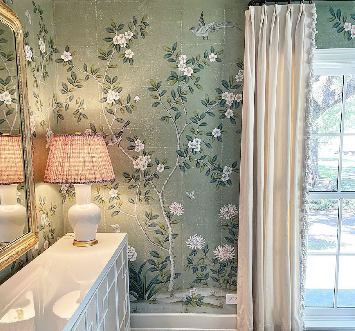 Close-up of chinoiserie hand-painted wallpaper 'Jade Garden' on antique-finish green tea paper, with white blossoms and birds, styled beside a white console table with a pleated lampshade and floor-length ivory drapes.