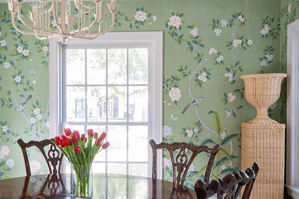 Chinoiserie hand-painted wallpaper 'Jade Garden' on green tea paper with antique finish, featuring white and pink blossoms and birds, styled in a traditional dining room with a round wooden table and classic carved chairs.