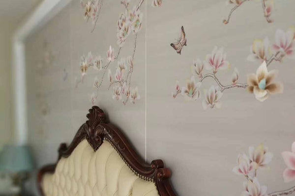 Hand-painted magnolia and butterfly mural with partial embroidery, installed as bedroom headboard backdrop on neutral-toned wallpaper
